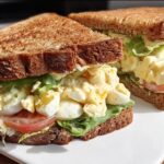 Two halves of a delicious restaurant-style egg salad sandwich with lettuce and tomato on whole wheat bread.