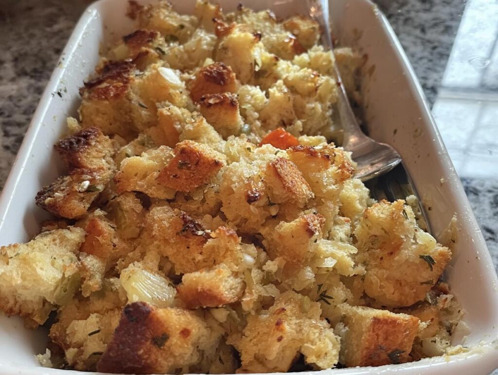 Close-up of a white baking dish filled with golden-brown restaurant-style stuffing, featuring cubes of bread and visible herbs.