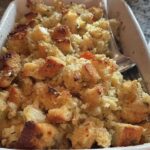 Close-up of a white baking dish filled with golden-brown restaurant-style stuffing, featuring cubes of bread and visible herbs.
