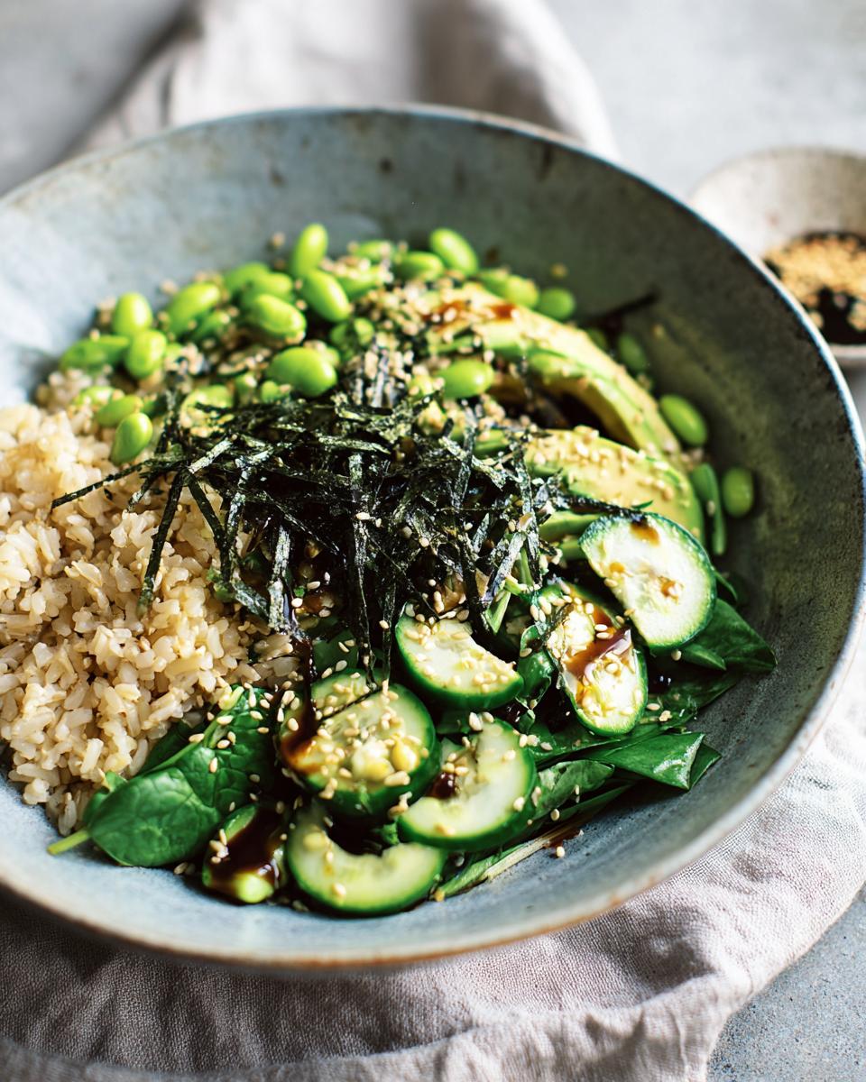 A vibrant rice bowl featuring brown rice, sliced avocado, edamame, cucumber, spinach, and nori, drizzled with sauce.