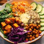 A vibrant and colorful rice bowl recipe featuring quinoa, roasted sweet potatoes, chickpeas, spinach, shredded carrots, cucumber, and red cabbage.