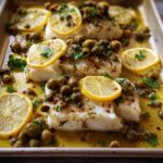 Close-up of baked white fish fillets topped with lemon slices, olives, capers, and herbs on a sheet pan.