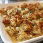 A close-up of a baked casserole dish filled with a savory stuffing recipe, topped with golden-brown bread cubes and fresh herbs.