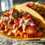 Close-up of two crispy fish tacos for Taco Tuesday, filled with fresh pico de gallo, creamy sauce, and shredded vegetables.
