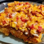 A close-up of a baked Taco Tuesday recipe casserole in a glass dish, topped with melted cheese, ground beef, corn, and tomatoes.