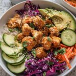 A close-up of a Bang Bang Chicken Bowl filled with rice, glazed chicken, avocado, cucumber, shredded carrots, and red cabbage.