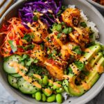 A vibrant bowl filled with rice, crispy salmon bites, avocado, cucumber, edamame, shredded carrots, and red cabbage, drizzled with Bang Bang sauce.