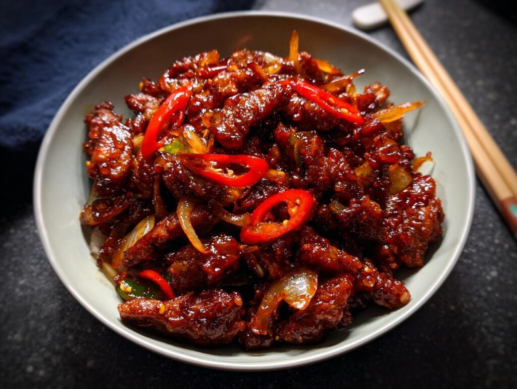 A close-up of Crispy Chilli Beef Rice, featuring tender beef strips coated in a glossy, spicy sauce with sliced red chillies and onions.