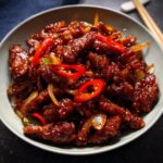 A close-up of Crispy Chilli Beef Rice, featuring tender beef strips coated in a glossy, spicy sauce with sliced red chillies and onions.