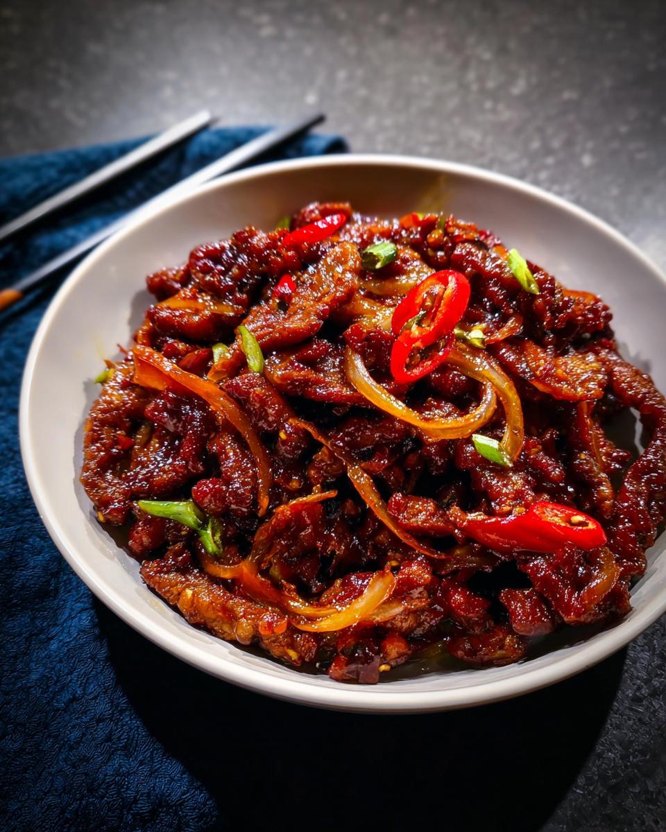 A close-up of a bowl filled with Crispy Chilli Beef Rice, featuring tender beef strips coated in a glossy sauce with sliced red chilies and onions.