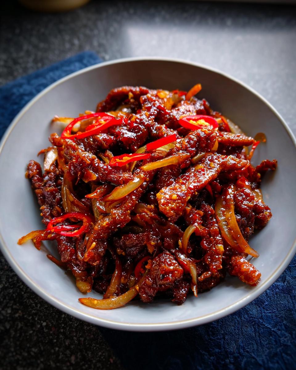 A close-up of a bowl filled with Crispy Chilli Beef Rice, featuring tender beef strips coated in a glossy sauce with sliced red chilies and onions.