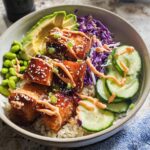 A vibrant Crispy Salmon and Rice Bowl featuring glazed salmon, edamame, avocado, cucumber, and red cabbage.