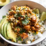 A vibrant Easy Street Corn Chicken Bowl topped with crumbled cheese, cilantro, and served with sliced avocado and lime wedges.