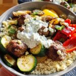 A close-up of a Greek Meatball Bowl featuring tender meatballs, couscous, grilled zucchini, bell peppers, and a creamy tzatziki sauce.
