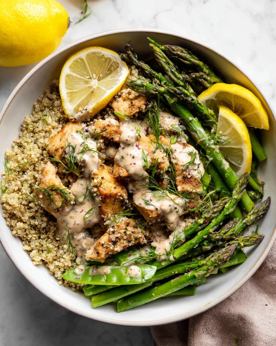 A Healthy Lemon Garlic Chicken Meal Prep Bowl with quinoa, asparagus, and snap peas, drizzled with sauce and garnished with lemon slices and dill.