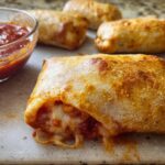 Close-up of a golden-brown HIGH PROTEIN Pepperoni Pizza Roll, oozing with melted cheese and served with a side of marinara sauce.