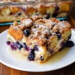 A delicious slice of Best Blueberry French Toast Casserole, topped with streusel and powdered sugar, on a white plate.