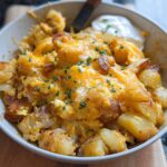 A close-up of a bowl filled with a Cheesy Potato Egg Scramble, topped with melted cheddar cheese and chives.