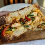 A close-up of a cut CHICKEN AVOCADO MELT SANDWICH with melted cheese, chicken, avocado, and red peppers.