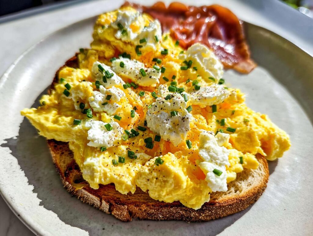 A slice of toast topped with fluffy scrambled eggs, dollops of ricotta cheese, and fresh chives.