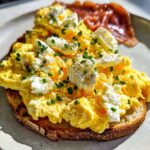 A slice of toast topped with fluffy scrambled eggs, dollops of ricotta cheese, and fresh chives.