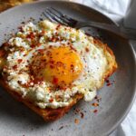 A close-up of crispy feta fried eggs served on toasted bread, sprinkled with chili flakes and herbs.