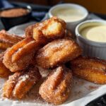 A pile of golden brown Easy Baked Churro Bites coated in cinnamon sugar, served with two dipping sauces.