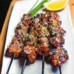 A plate of Easy Japanese Chicken Yakitori Skewers, glazed and sprinkled with sesame seeds and chives, served with a lemon wedge.
