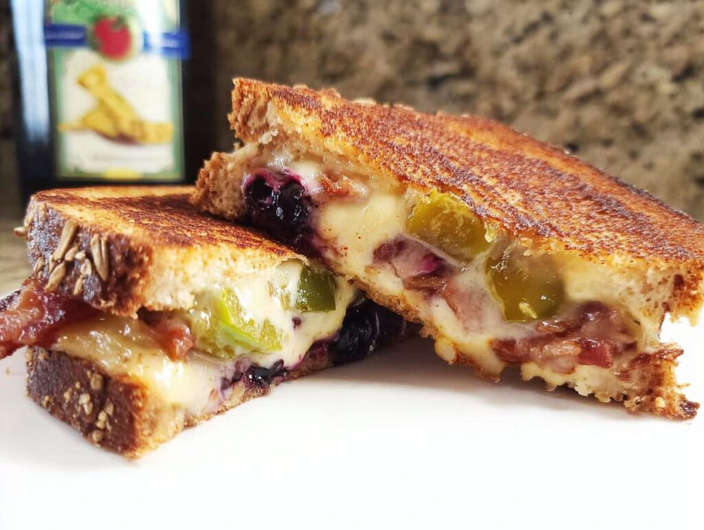 A close-up of a Gourmet Blueberry Grilled Cheese sandwich, cut in half, showing melted cheese, blueberries, and bacon.