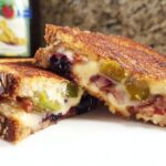 A close-up of a Gourmet Blueberry Grilled Cheese sandwich, cut in half, showing melted cheese, blueberries, and bacon.