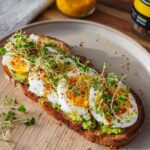 A high-protein breakfast toast topped with mashed avocado, poached eggs, and sprouts, sprinkled with chili flakes.