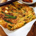 A freshly cooked, golden-brown Korean pancake (Pajeon) with visible green onions and a side of dipping sauce.