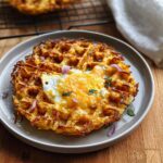 A golden-brown savory hashbrown chaffle topped with a fried egg and melted cheese, garnished with red onion and parsley.