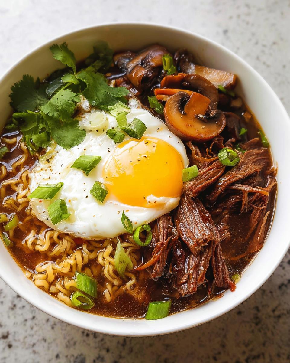 A bowl of Slow Cooker Beef Ramen Noodles topped with a fried egg, sliced mushrooms, cilantro, and green onions.