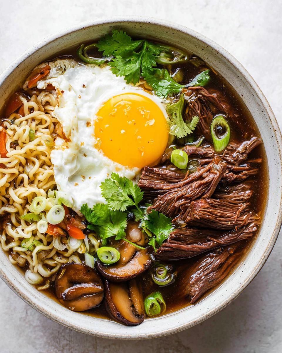 A close-up of a bowl of Slow Cooker Beef Ramen Noodles, topped with a fried egg, shredded beef, mushrooms, and cilantro.