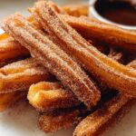 A close-up of a stack of golden-brown Spanish churro pancakes coated in cinnamon sugar, with a small bowl of chocolate dip in the background.