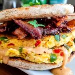 A close-up of a stacked Spicy Chipotle Egg Breakfast Sandwich with crispy bacon and creamy sauce.