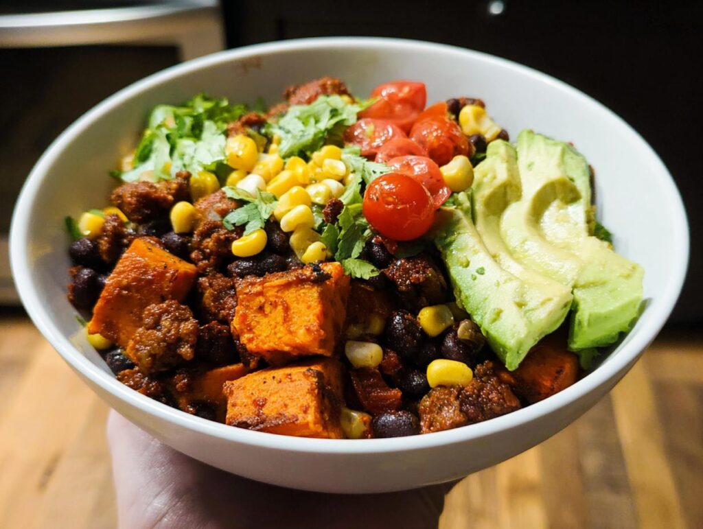 A vibrant Sweet Potato Taco Bowl filled with roasted sweet potatoes, black beans, corn, cherry tomatoes, avocado slices, and shredded lettuce.