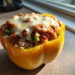 A close-up of a yellow bell pepper stuffed with rice, ground meat, and green peppers, topped with melted cheese.