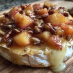Close-up of baked brie topped with warm peaches and toasted pecans, with melted cheese oozing out.