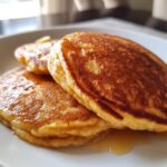 Close-up of a stack of golden-brown banana oat pancakes drizzled with syrup on a white plate.