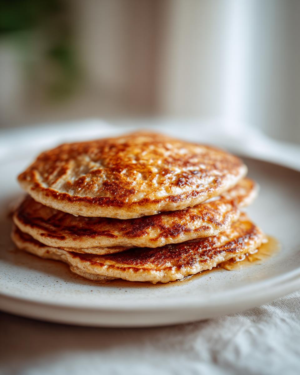 A close-up shot of a stack of three golden-brown banana oat pancakes on a plate, drizzled with syrup.