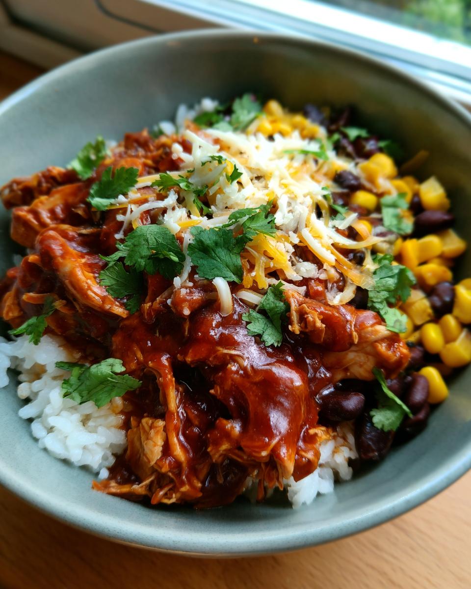 A close-up of a BBQ chicken bowl with rice, shredded chicken in BBQ sauce, black beans, corn, cheese, and cilantro.