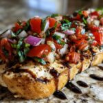 A close-up of a grilled chicken breast topped with fresh bruschetta mixture of tomatoes, red onion, and basil, drizzled with balsamic glaze.