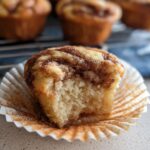 A close-up of a Cinnamon Roll Protein Muffin with a bite taken out, showing the fluffy interior and cinnamon swirl.