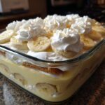A glass dish filled with layers of classic banana pudding, sliced bananas, vanilla wafers, and topped with homemade whipped cream.
