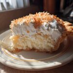 A generous slice of coconut cream pie, topped with whipped cream and toasted coconut flakes, on a decorative plate.
