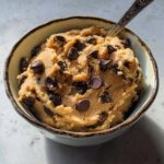 A bowl filled with delicious protein cookie dough loaded with chocolate chips, with a spoon inside.