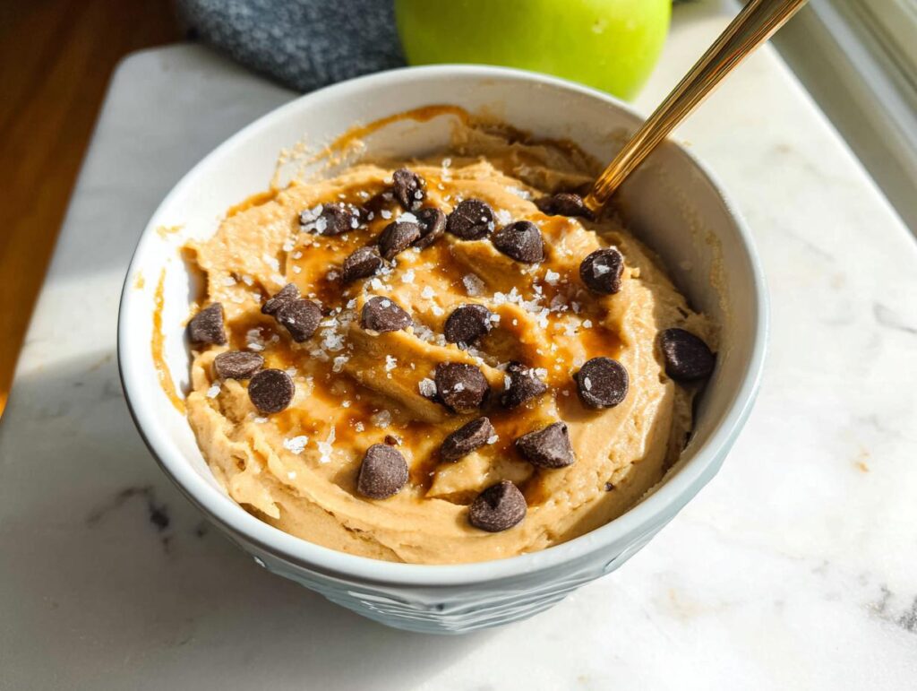 A bowl of Greek Yogurt Cookie Dough topped with chocolate chips, caramel drizzle, and sea salt, with a spoon.
