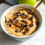 A bowl of Greek Yogurt Cookie Dough topped with chocolate chips, caramel drizzle, and sea salt, with a spoon.